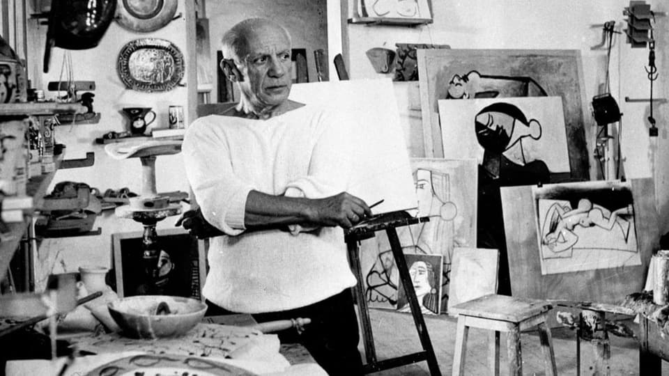 Der Künstler Pablo Picasso steht in seinem Atelier umgeben von Bildern und anderem Kram.