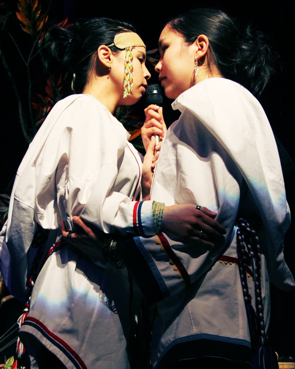 Zwei Frauen in traditioneller Kleidung stehen eng beieinander und singen in ein Mikrofon.