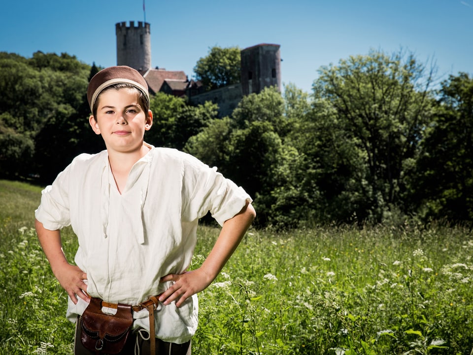 Junge in mittelalterlicher Kleidung auf einer Wiese mit einer Burg im Hintergrund
