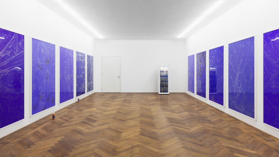 Ein Museumsraum mit Parkett. An den Wänden hängen blaue Bilder. Am Bodens stehen Flaschen.