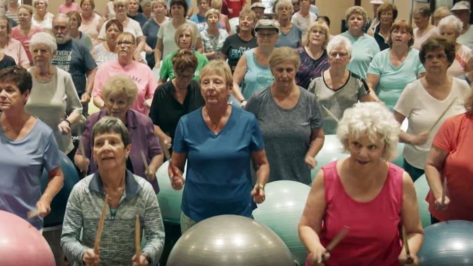 Eine grosse Gruppe von Senioren und Seniorinnen, welche mit Stöcken auf Gymnastikbällen trommeln. 