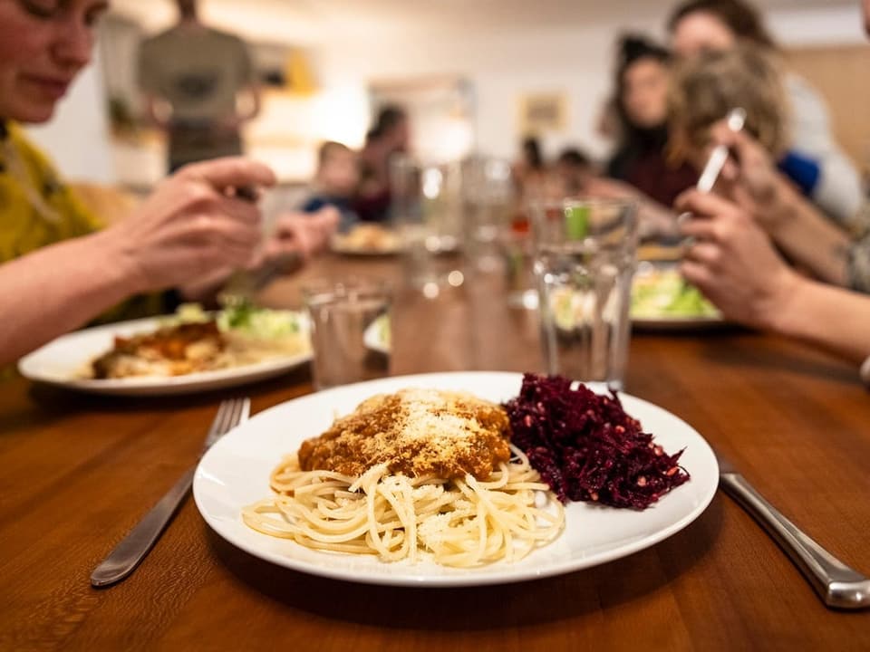 Ein Tisch, im Vordergrund ein Teller mit Spaghetti und Tomatensauce, im Hintergrund viele Personen (verschwommen).