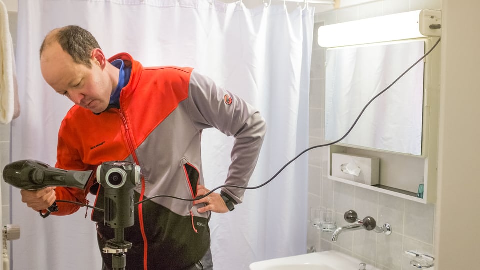 Ein Mann benutzt in einer Toilette den Heissluftföhn, um eine Kamera zu erwärmen. 