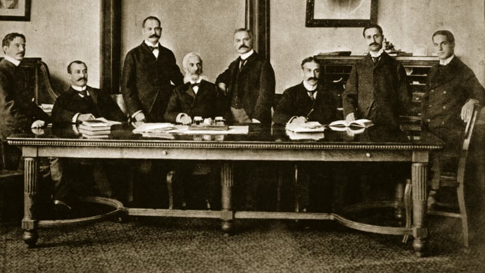 Schwarz-weiss Bild von gut gekleideten Männer um einen Tisch. 