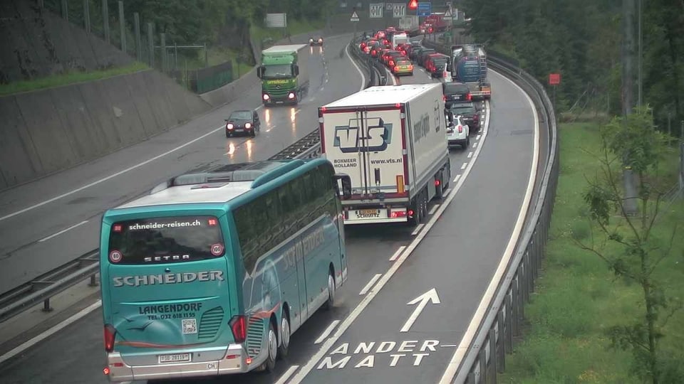 Stau vor dem Gotthardtunnel. Man sieht vorne im Bild einen grünlichen Car und einen weisslichen Lastwagen. Vor ihnen stauen sich mehrere Autos. Es ist bewölkt, die Fahrbahn nass.