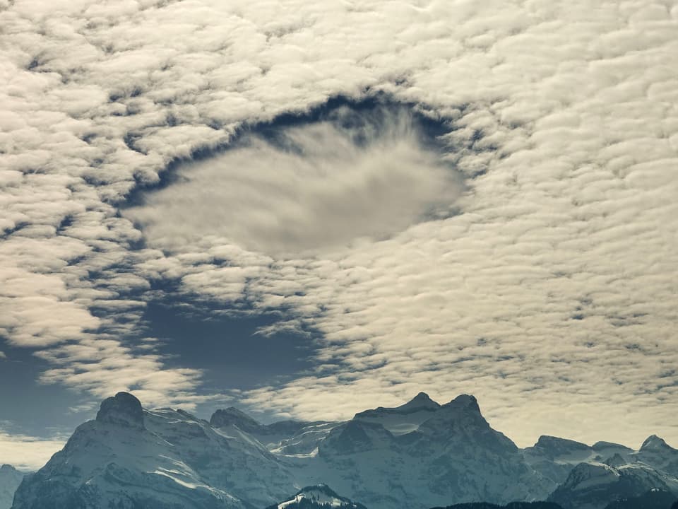 Am Himmel hat es ein grosses Wolkenfeld. In der Mitte hat es ein Loch mit einer Wolke in einer wenig tieferen Luftschicht. Am Horizont sind verschneite Berge zu sehen.