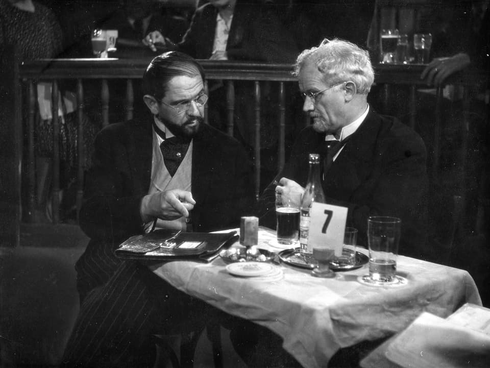 Zwei ältere Männer sitzen an einem Tisch in einem Restaurant. Auf dem Tisch liegt eine Aktenmappe. Der linke Mann zeigt mit dem Finger auf die Mappe.