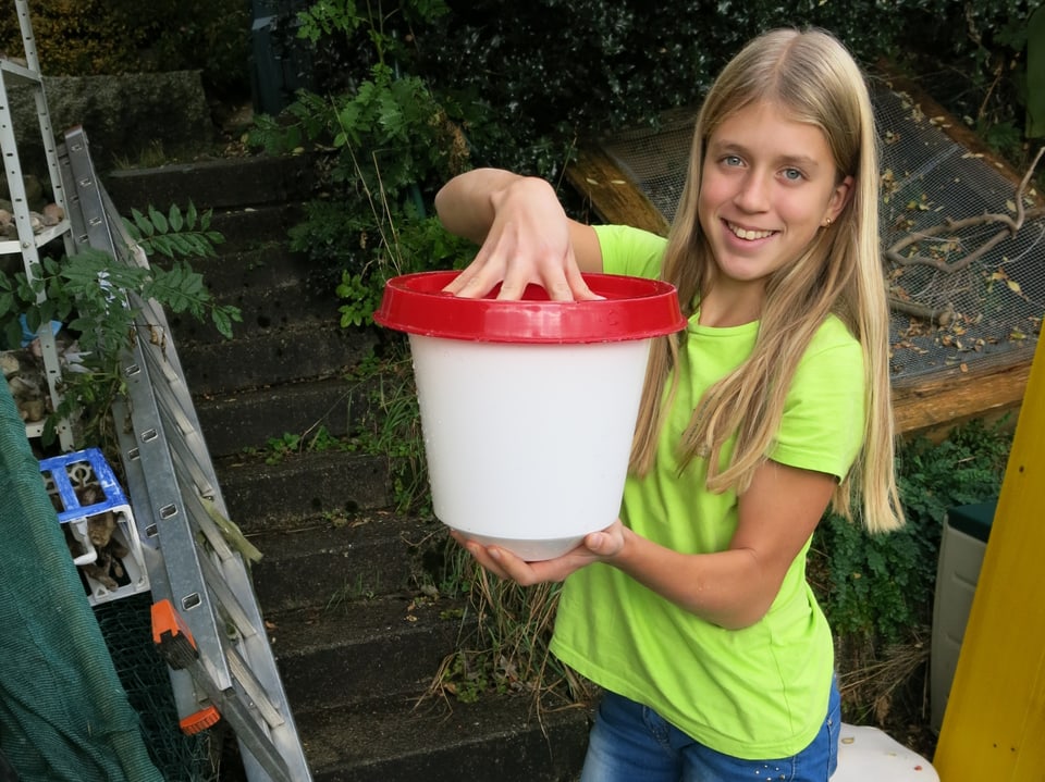 Elena hält den weissen Wasserbehälter mit rotem Deckel in beiden Händen, eine Hand oben, die andere unten.