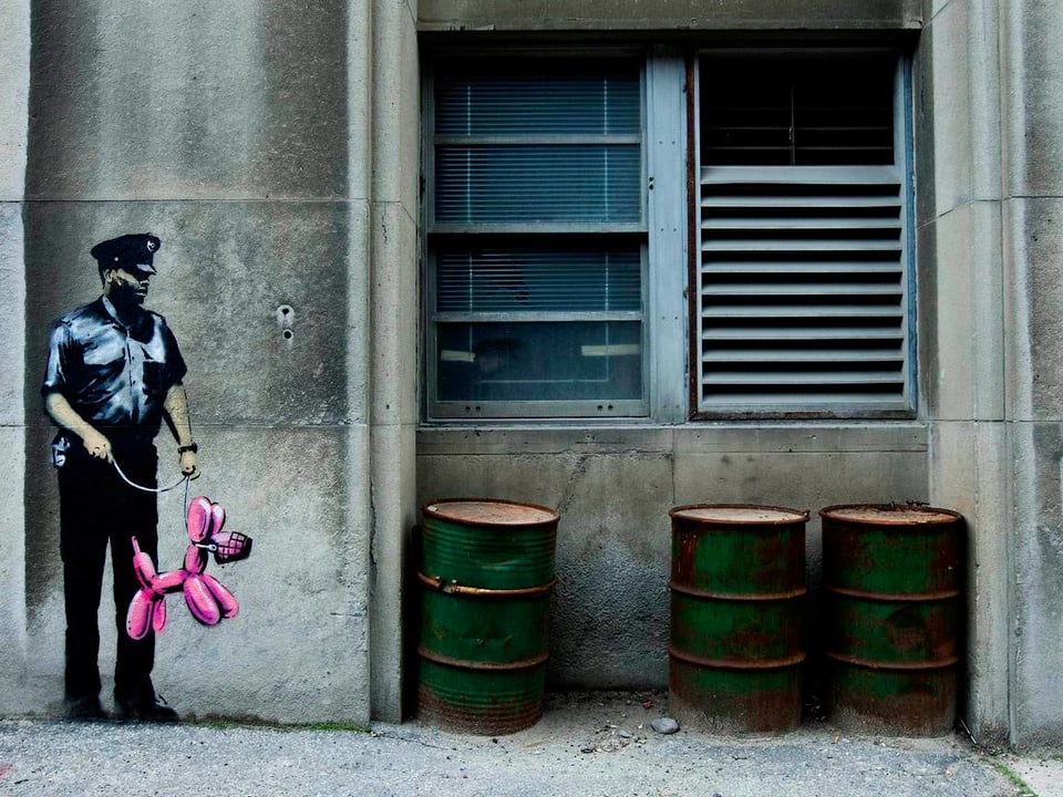 Auf einer grauen Wand ist ein Polizist aufgemalt, der einen pinkfarbenen Ballonhund an der Leine hält.