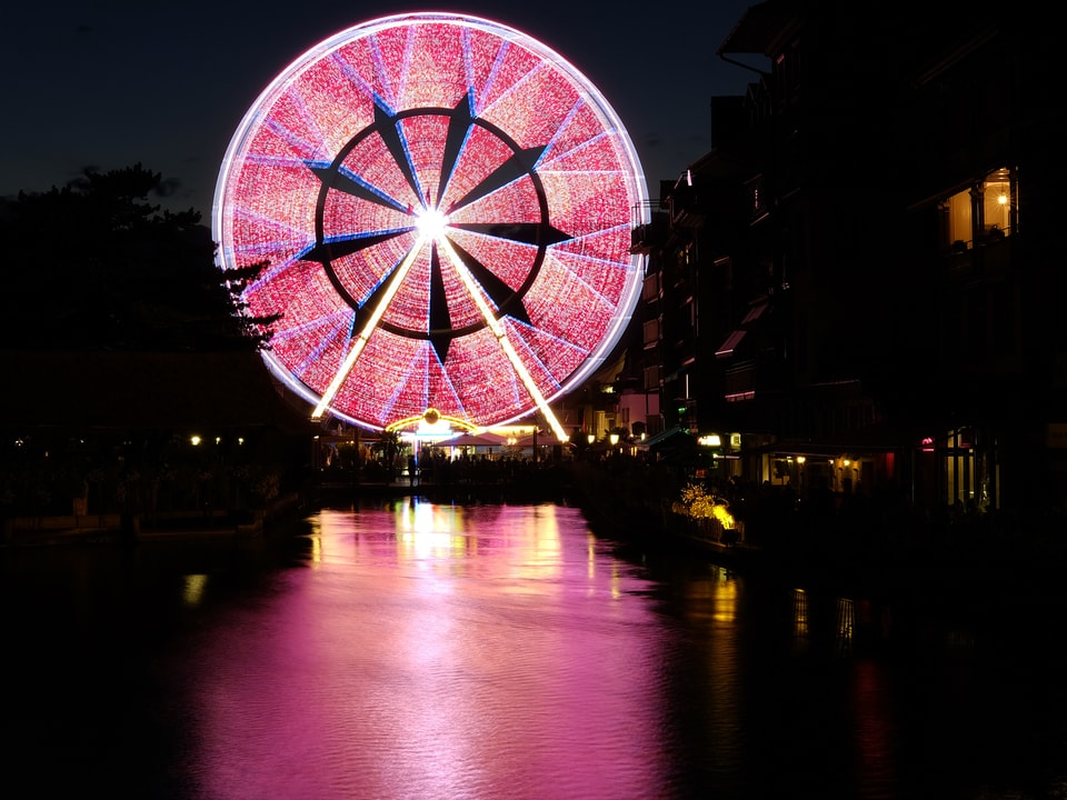 Rosarot leuchtendes Riesenrad, dessen Lichter sich im Fluss spiegeln.