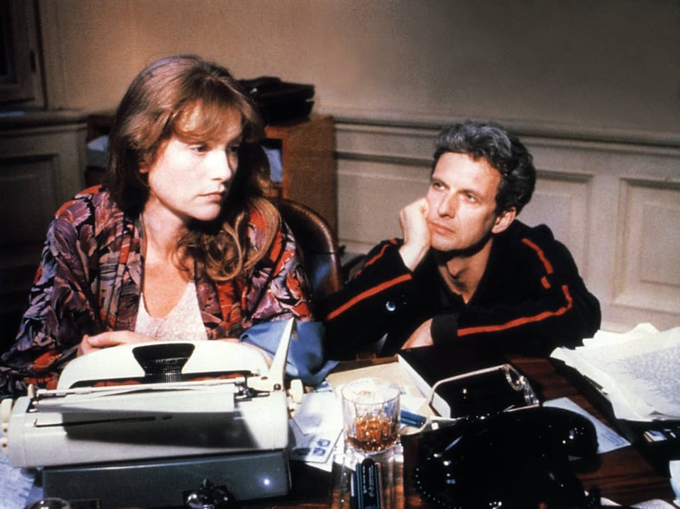 Film still, on the left a woman with brown hair in a dressing gown sits at a typewriter, on the right a man looks at her