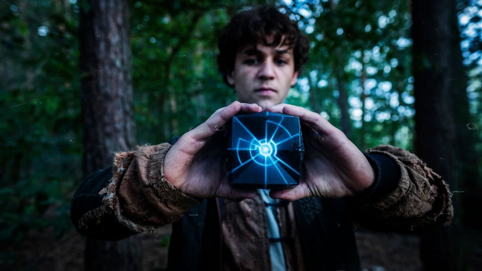 Ein Junge im Wald hält einen backsteinähnlichen Stein in der Hand, der blau leuchtet.