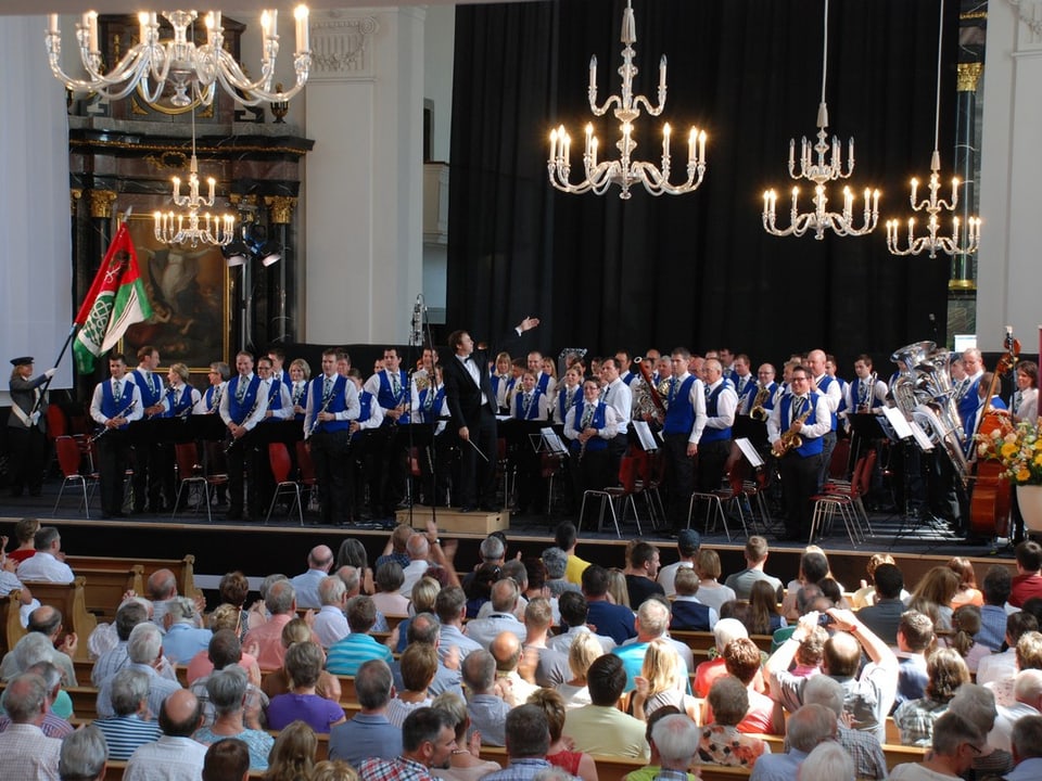 Eine Blasmusikformation in weissen Hemden und blauem Gilet steht auf einer Bühne in einer Kirche.