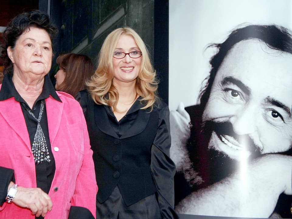 Die Witwe Nicoletta Mantovani in pinker Jacke und die in Schwarz gekleidete Schwester Gabriella Pavarotti stehen während einer Ausstellung neben einer grossen Schwarz-Weiss-Fotografie von Sänger Pavarotti. 