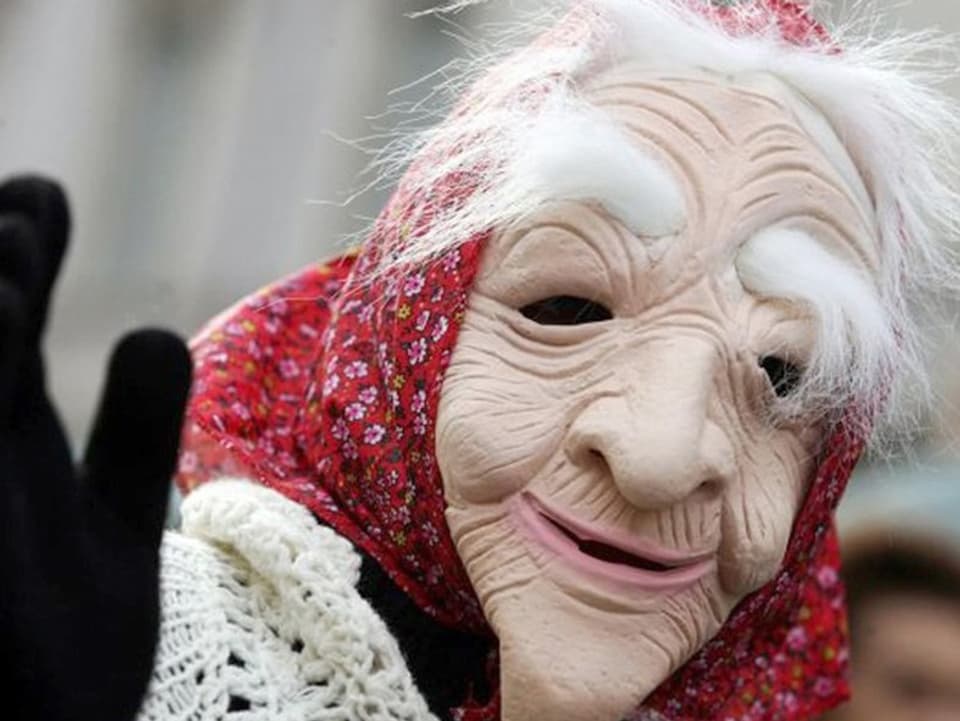 Jemand trägt die Befana-Maske. Sie zeigt das Gesicht einer alten Frau mit Kopftuch und weissen Haaren.