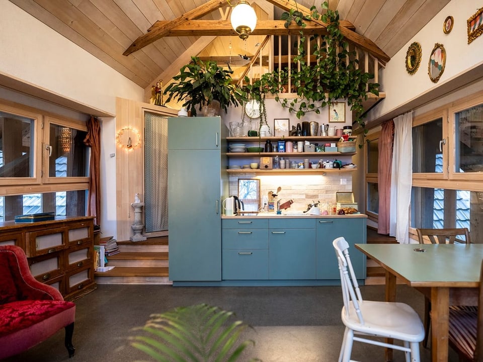 Ein Raum mit einer kleinen Kochnische, einem Tisch mit Stühlen und einer Gallerie, die mit Pflanzen behangen ist.