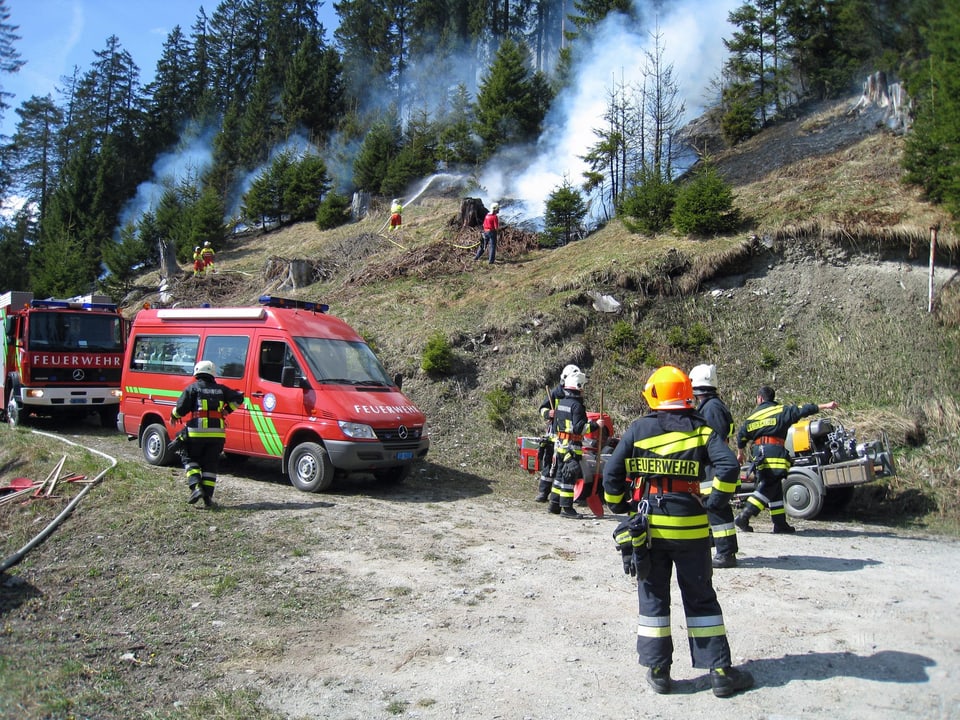 Feuerwehr an einem Berghang mit Bäumen. Es sind mehrere Rauchfahnen am Hang erkennbar.