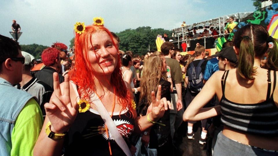 Links junge Frau mit rotem Haar und gelben Blumen im Haar mit Peacezeichen, dahinter Menschenmasse, hinten eine Bühne.