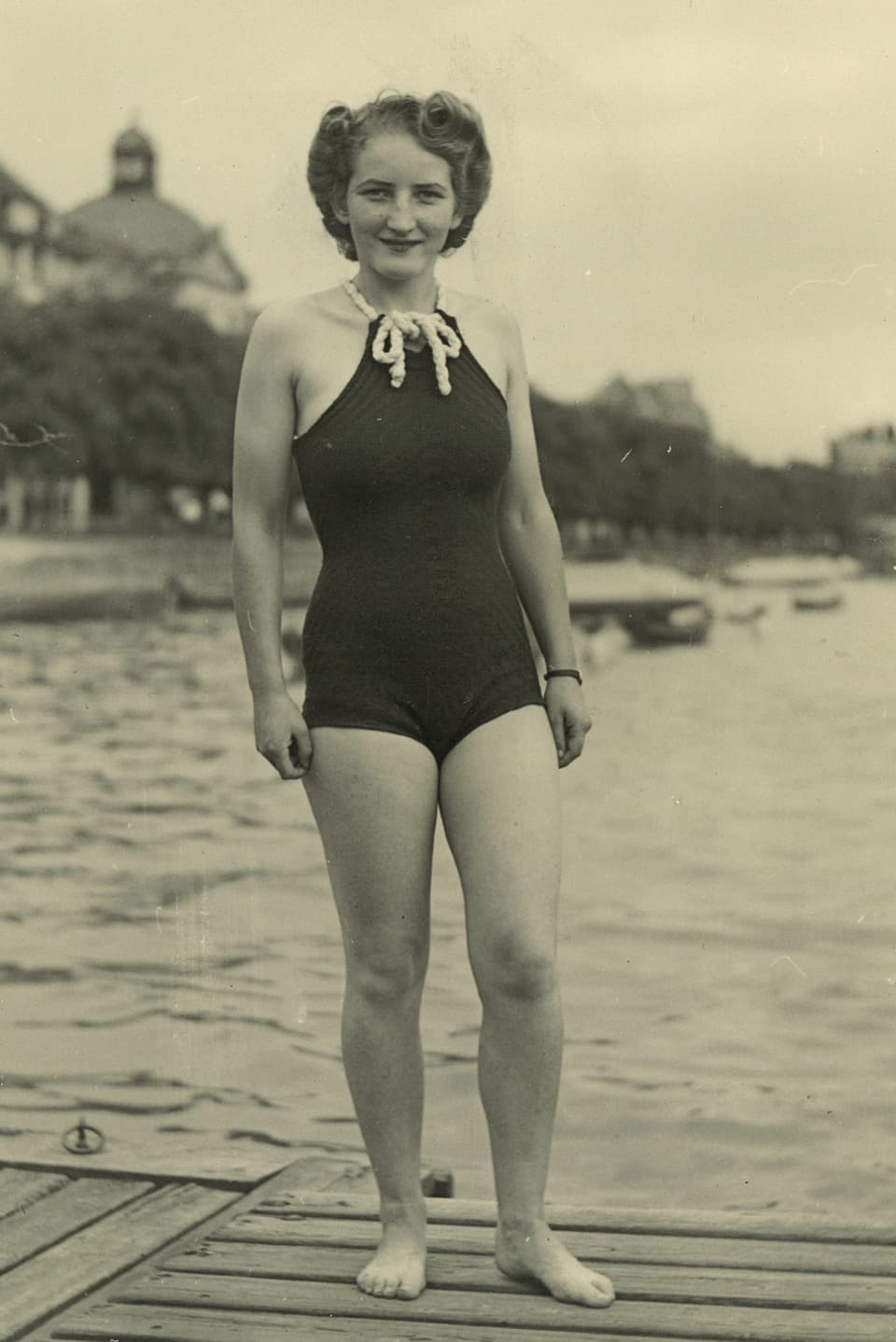 Schwarz-Weiss-Fotografie mit einer jungen Frau im Badeanzug auf einem Steg am Seeufer.