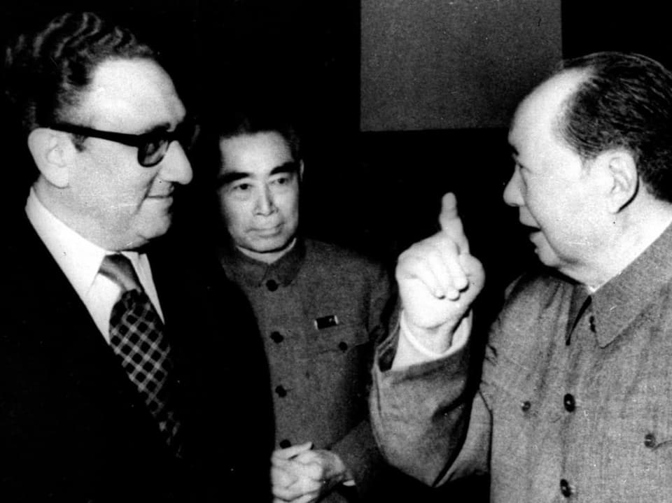 Henry Kissinger steht neben Mao Zedong, der seinen rechten Zeigefinger hochhält.