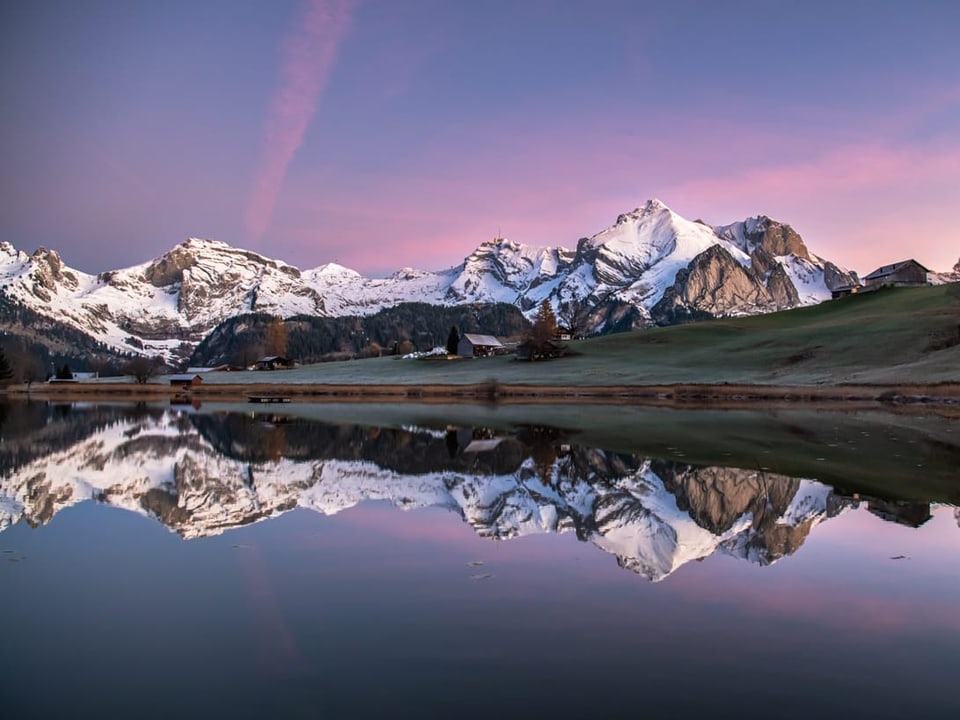 Berge und Landschaft wird in Bergsee gespiegelt. Dazu Himmel mit einigen rosaroten Wolken.
