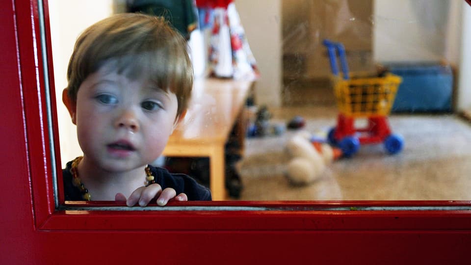 Ein Kind schaut durch die Glasscheibe einer Tür in einer Berner Kindertagesstätte