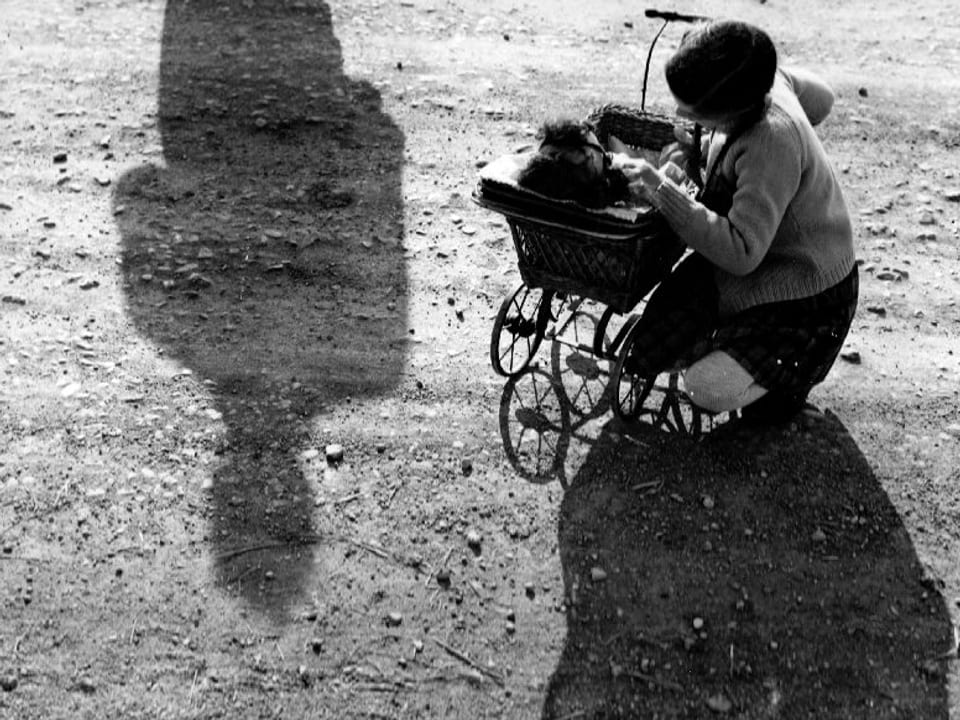 Eine Frau kniet neben einem Kinderwagen, in welchem ein Kleinkind liegt. Hinter ihr auf dem Boden zeichnet sich der Schatten eines Mannes ab.