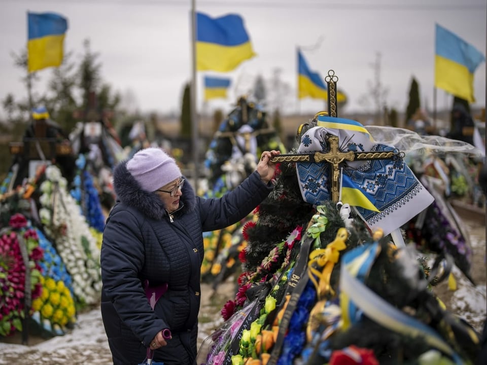 Eine Frau steht an einem geschmückten Grab und hält das Grabkreuz fest. Viele Ukraine-Flaggen.
