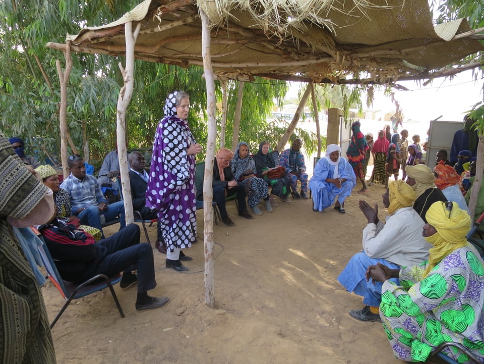 Christine Beerli spricht in einem bunten afrikanischen Gewand vor einer Dorfgemeinschaft unter einem einfachen Unterstand.