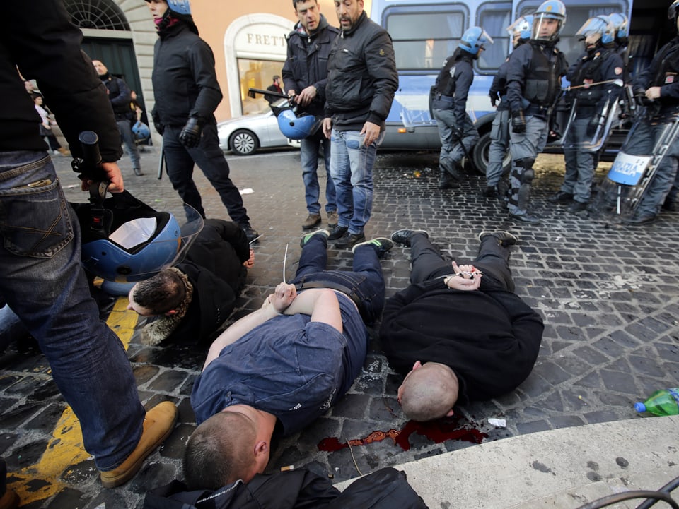 Zwei gefesselte Hooligans liegen mit dem Gesicht auf dem Boden von Polizisten umzingelt.