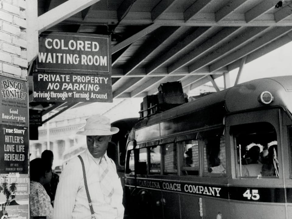 Ein schwarzer Mann steht an einer Bus-Station. Über seinem Kopf ist ein Schild mit der Aufschrift: «Colored Waiting Room».