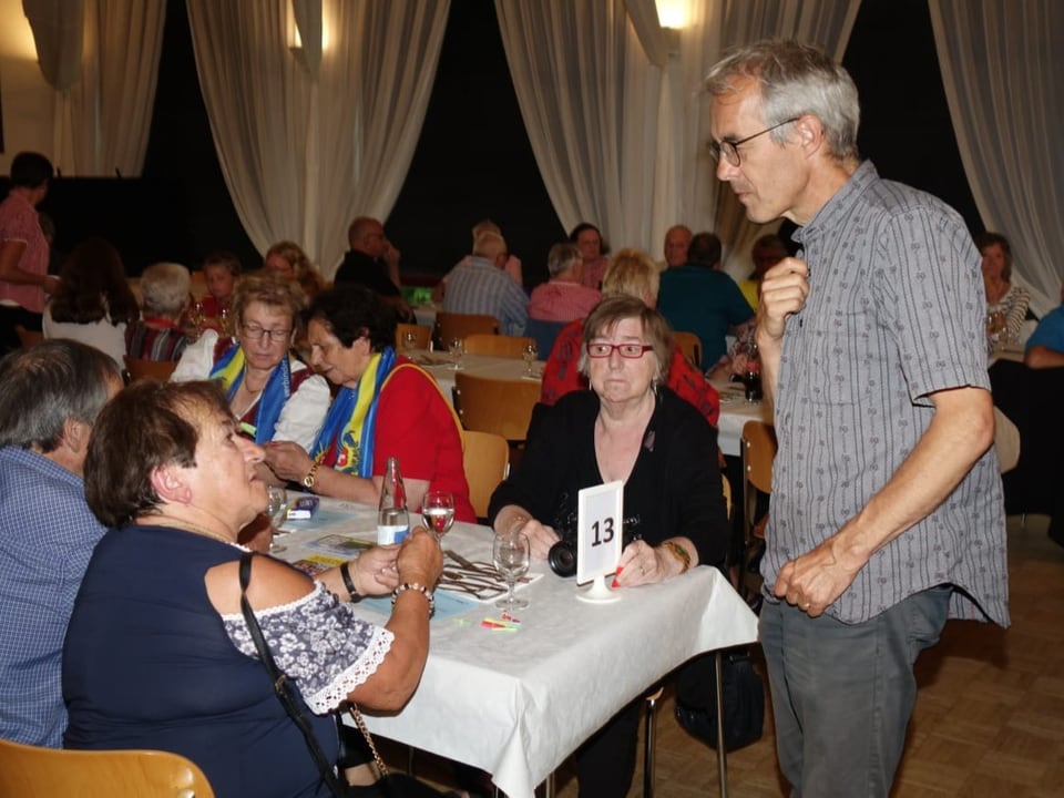 Ältere Menschen sitzen in einem Festsaal am Tisch, ein stehender Mann im Sennenhemd hört ihnen zu