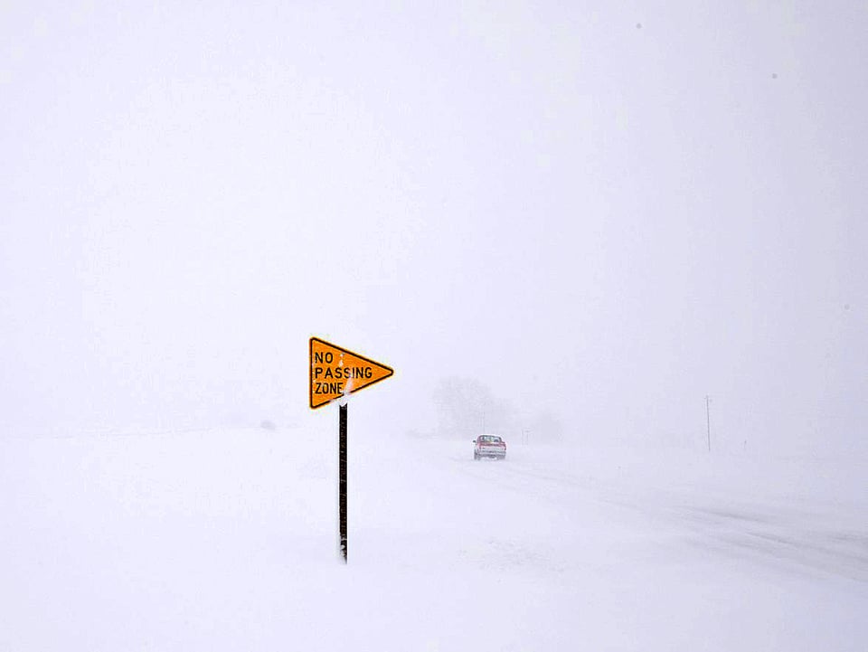 Ein einsames gelbes Strassenschild im undurchdringbaren Weiss eines Schneesturms. Weiter hinten im Bild die Andeutung eines Autos.