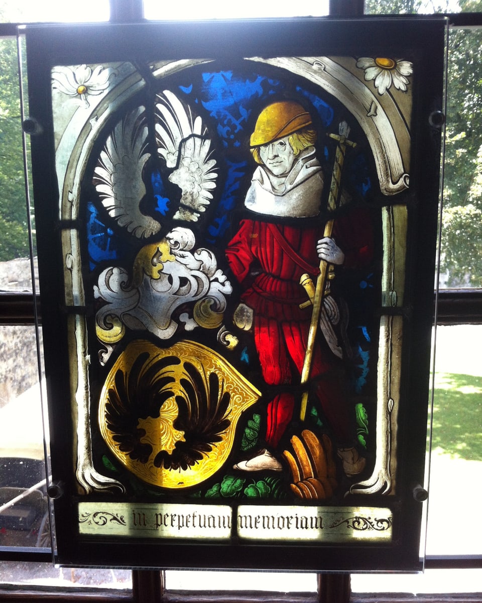 Ein schön verziertes Glasfenster. Darauf ist eine heilige Figur und ein Wappen zu erkennen.
