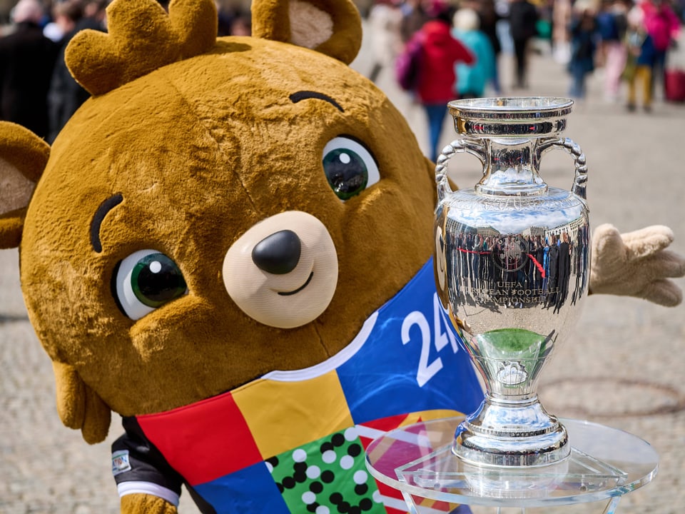 Maskottchen der EM 2024 mit einem Fussballtrikot hält einen Pokal.