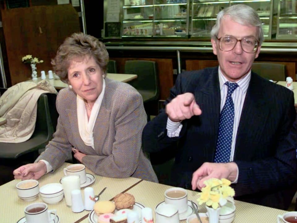 Eine Frau mit grauem Blazer sitzt mit einem Mann an einem Tisch. Der Mann trägt eine blaue Krawatte mit weissen Punkten. Auf dem Tisch stehen Kaffee und Süssgebäck.