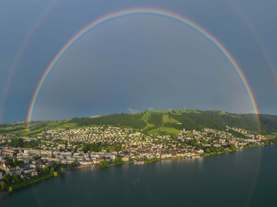 Regenbogen über Landschaft mit See, Stadt am Ufer und grünem Berg