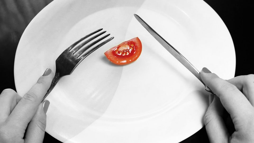 Eine einsame Tomate auf einem Teller, darüber Messer und Gabel in den Händen einer Frau.
