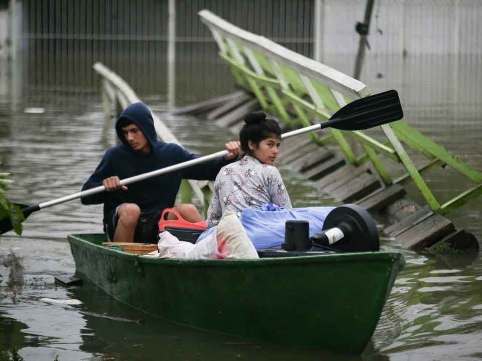 Zwei Personen in einem grünen Boot auf überflutetem Gebiet neben einer versunkenen Brücke.