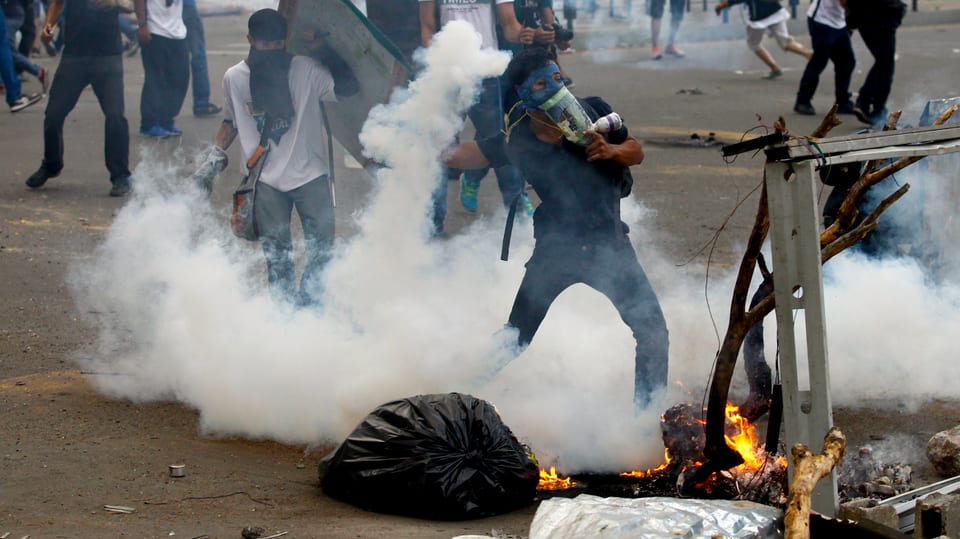 Vermummte Demonstranten in Venezuela. Einer wirft eine Rauchbombe.