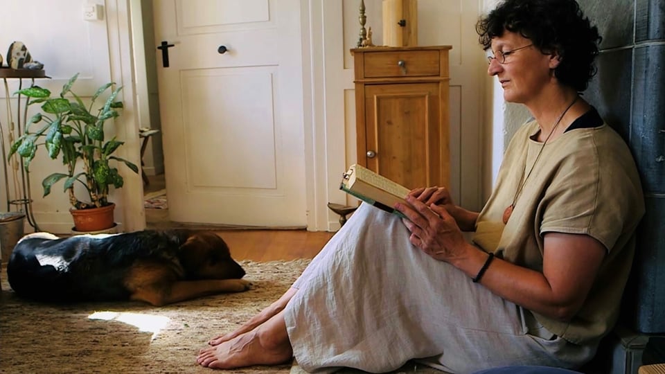 Eine Frau sitzt am Boden und liest ein Buch. Neben ihr liegt ein Hund.