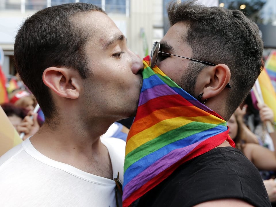 Zwei Männer küssen sich durch ein Regebogen-Tuch, das der eine um den Hals und über den Mund trägt.