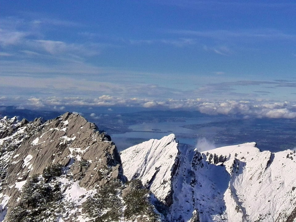 Vom schneebedeckten Gipfel des Mattstocks aus sieht man in der Ferne den Zürichsee. Der Himmel ist blau, im Flachland hat es viele tiefe Wolken.