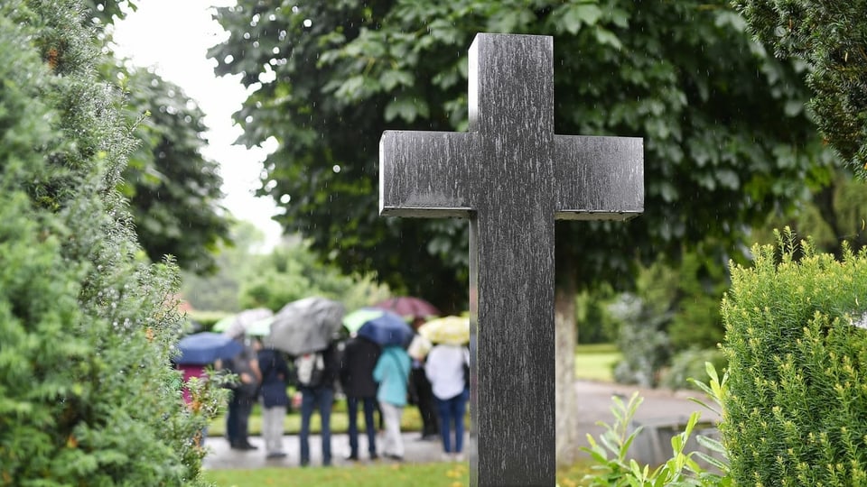 Leute mit Regenschirmen auf einem Friedhof, davor ein Kreuz.