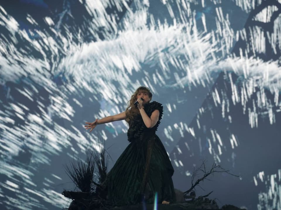 Sängerin in einem dunklen Kleid performt auf einer Bühne vor einem abstrakten, hellen Hintergrund.