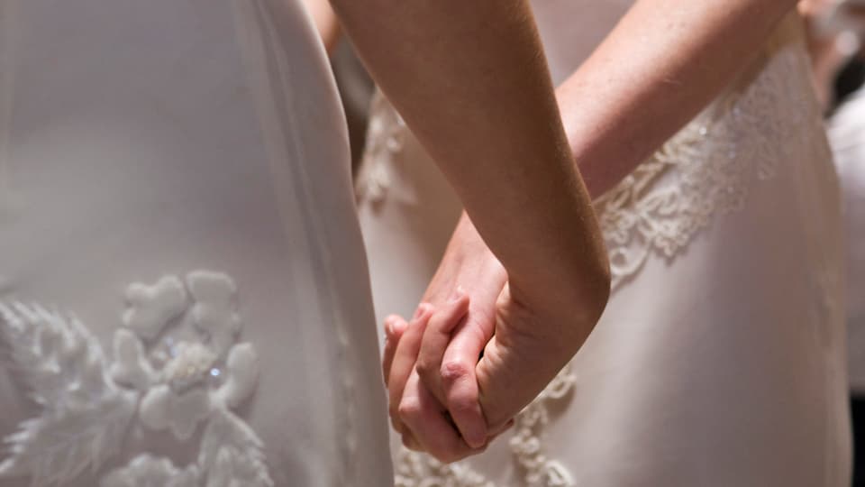 Zwei Frauen im Brautkleid halten sich an den Händen.