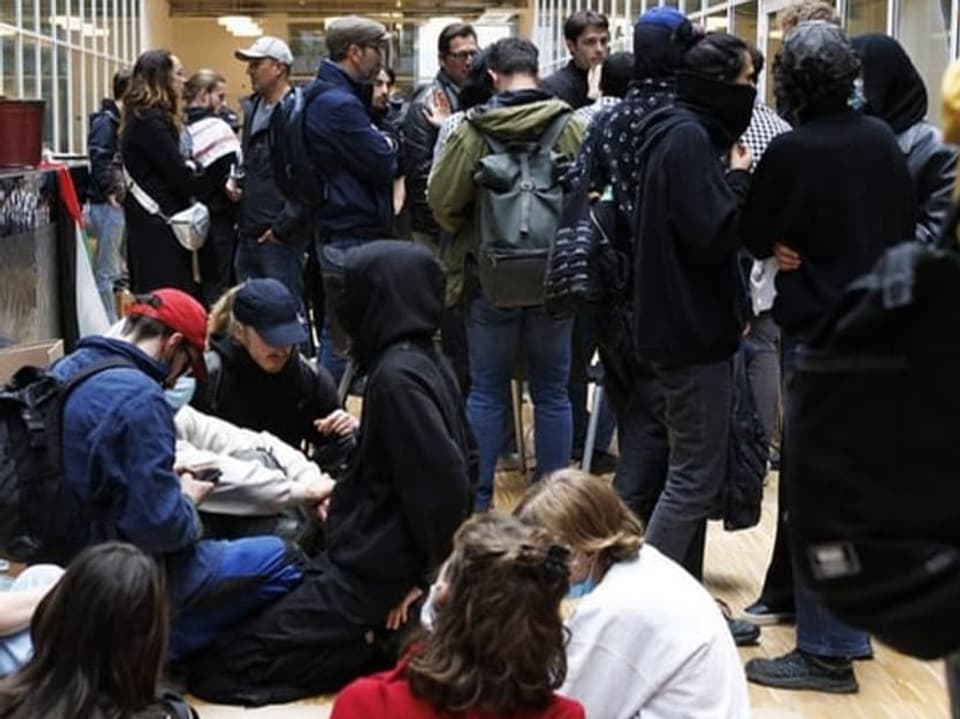 Gruppe von Menschen in einer belebten Korridorhalle, einige sitzen auf dem Boden, andere stehen.