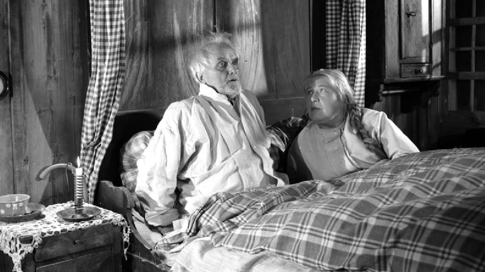 Ein älteres Ehepaar im Bett. Er sitzt aufgeschreckt im Bett, sie blickt besorgt zu ihm hoch.