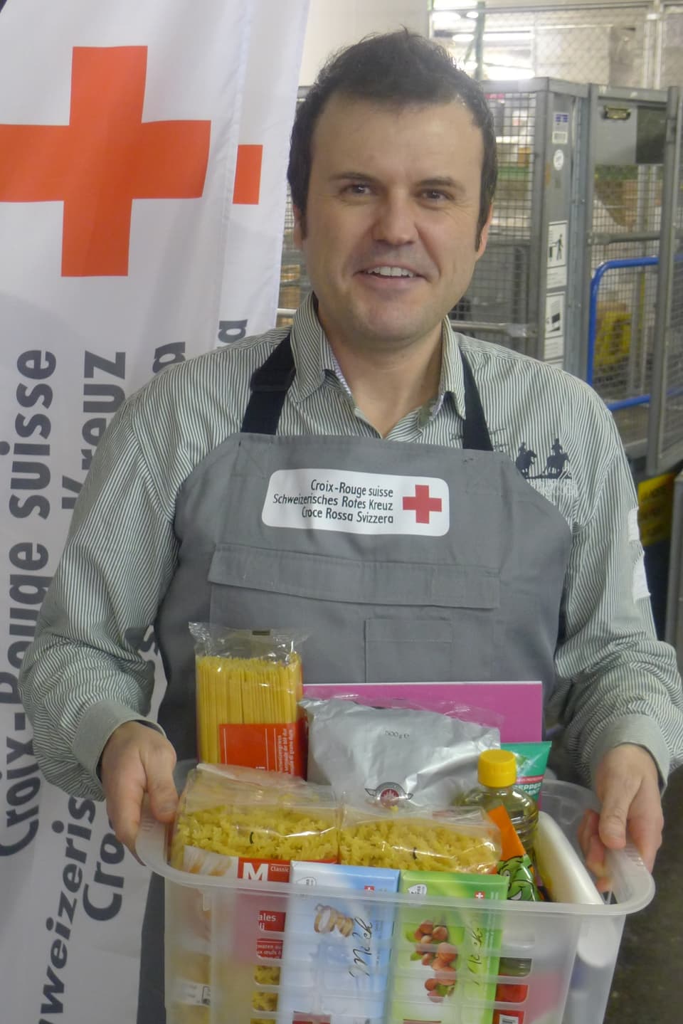 Joël Gilgen im Verteilzentrum des Schweizerischen Roten Kreuzes in Wabern mit einen Korb voll verschiedener Nahrungsmittel.