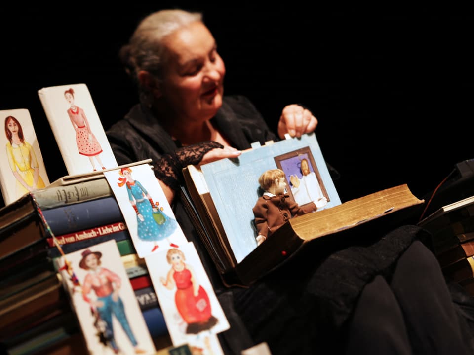 Margrit Gysin hält ein Buch, eine Puppe sitzt auf der aufgeschlagenen Seite. 